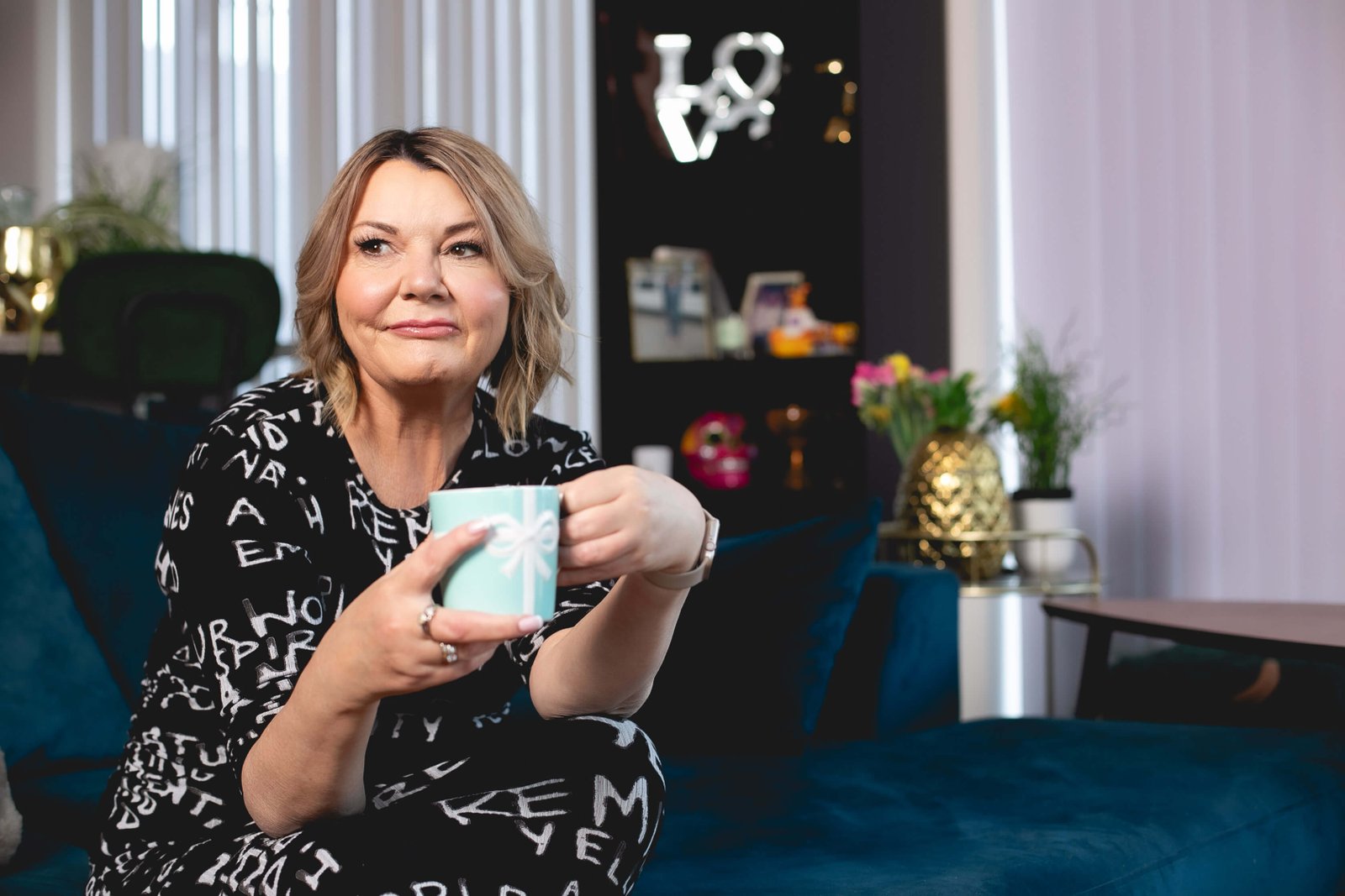 Nicky Wake wearing a black patterned dress sitting on a sofa holding a mug after quitting alcohol and embracing sober life - midlife sobriety