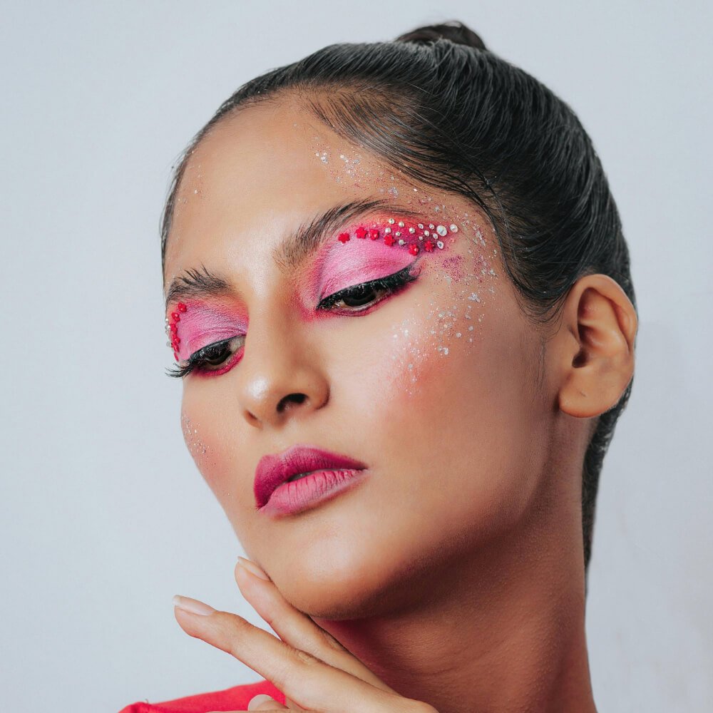 A close-up of a woman's face featuring dramatic pink and red gem makeup with glitter