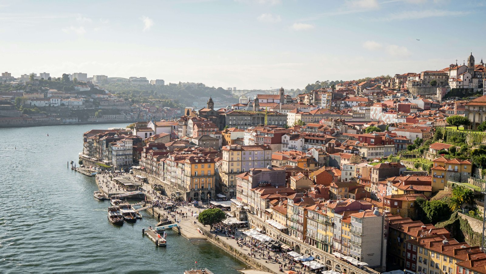 Aerial view of Porto’s Ribeira district in Portugal with colourful buildings and red-tiled roofs lining the Douro River, boats docked along the waterfront, and people walking along the lively riverside promenade.