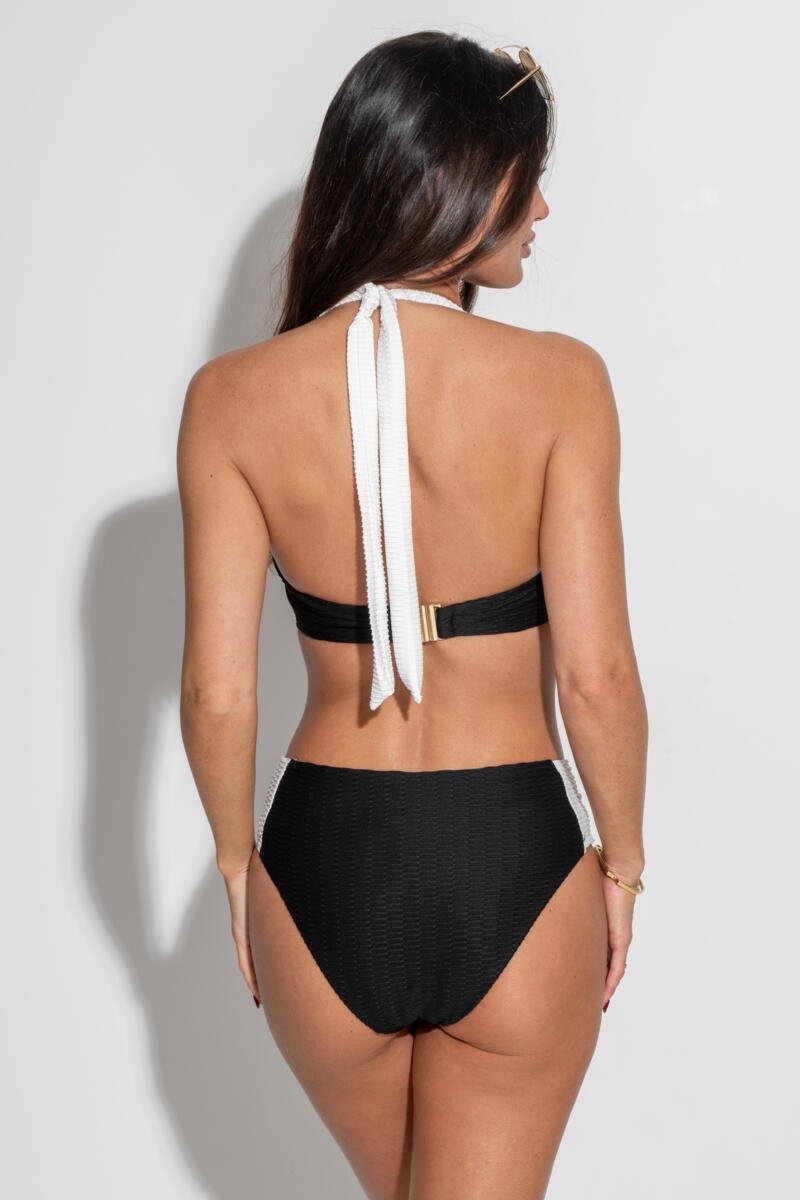 Rear view of a model posing against a white wall in the Pour Moi Acapulco textured bikini for big boobs. The black underwired bikini top features white halter straps tied at the neck and a gold clasp back closure, paired with matching black textured high-waisted bottoms with white side panels.