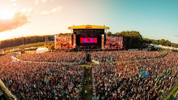 A wide, high-angle view of a massive, dense crowd packed into an outdoor field facing the Main Stage at the Leeds Festival, under a bright afternoon sky.
