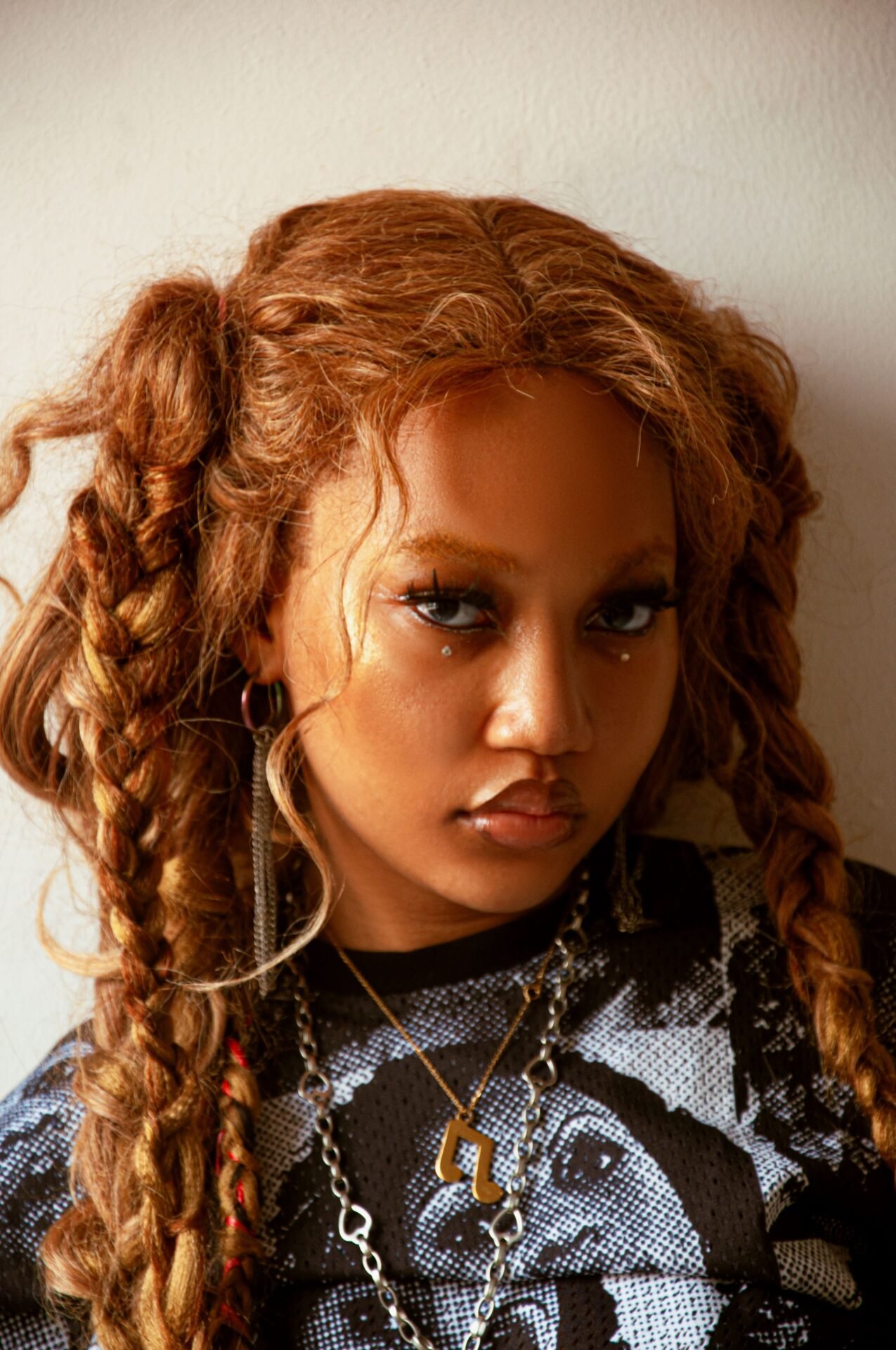 A young person with long, braided hair, wearing a patterned shirt and various accessories, poses against a plain background