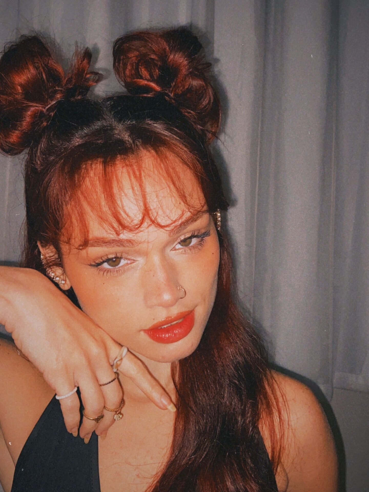 A woman with two buns in her hair poses for the camera, resting her chin on her hand, and has a serious expression with glossy red lips
