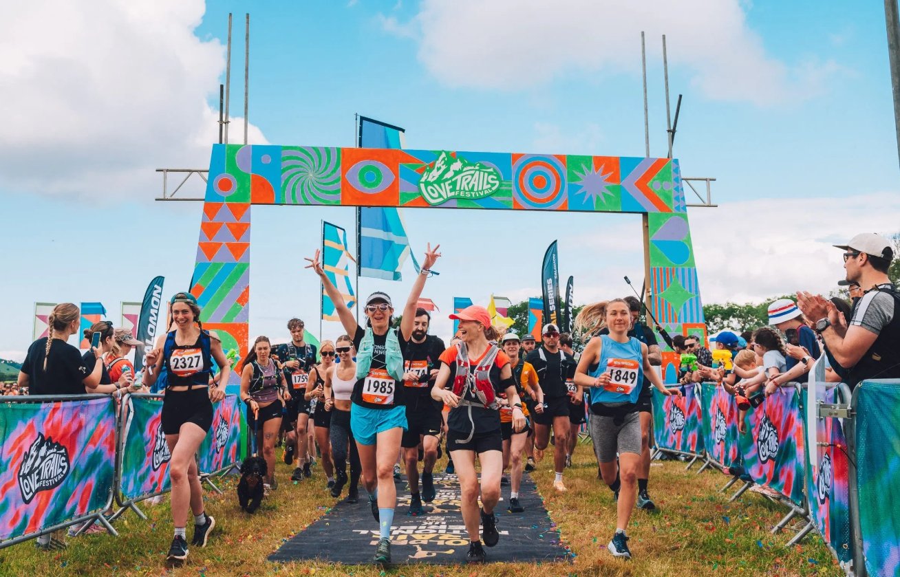 A group of enthusiastic trail runners cross a finish line under a vibrant, colorful geometric archway. The runner in the center cheers with both arms raised, while spectators clap from behind barriers at the Love Trails Festival.