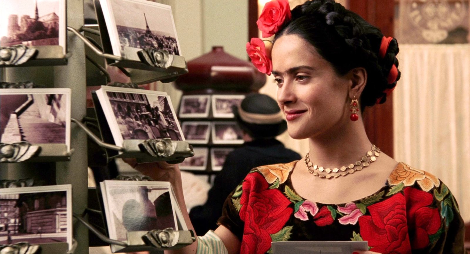Salma Hayek as Frida Kahlo, wearing a floral dress and admiring a rack of postcards in a scene from 'Frida'.