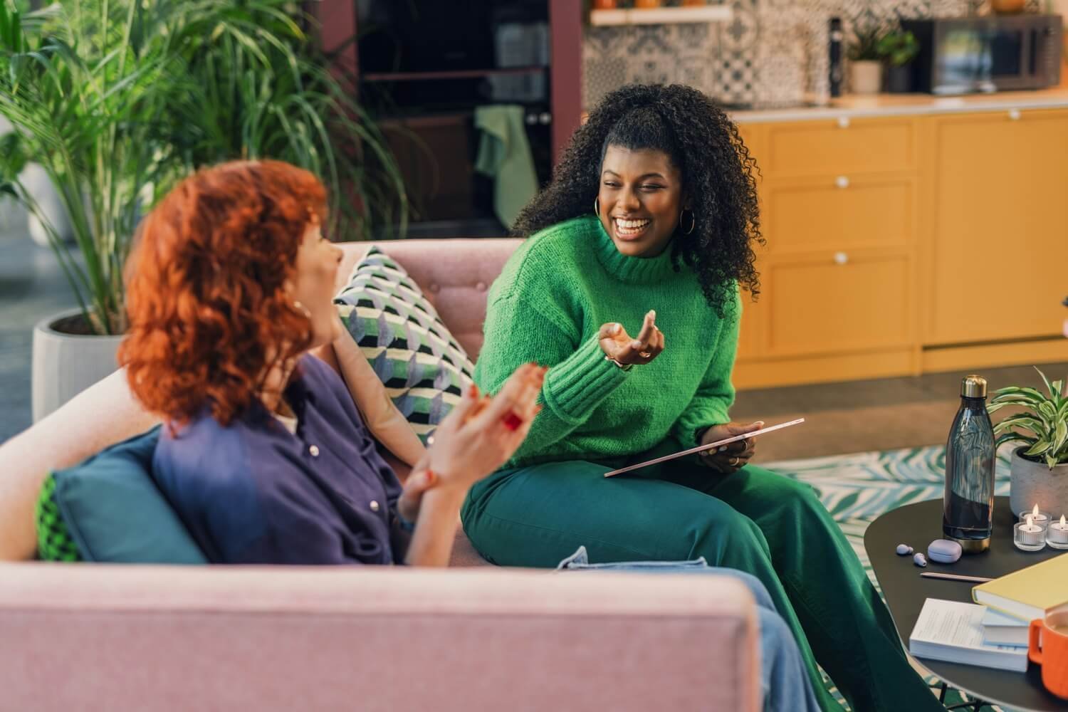 Two women laughing and chatting on a pink couch in a cosy room