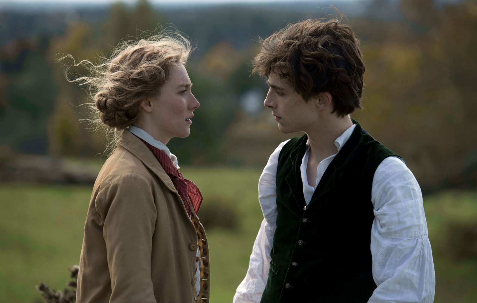Saoirse Ronan as Jo March and Timothée Chalamet as Laurie, both wearing period outfits and looking at each other in the middle of an open field in a scene from 'Little Women'.
