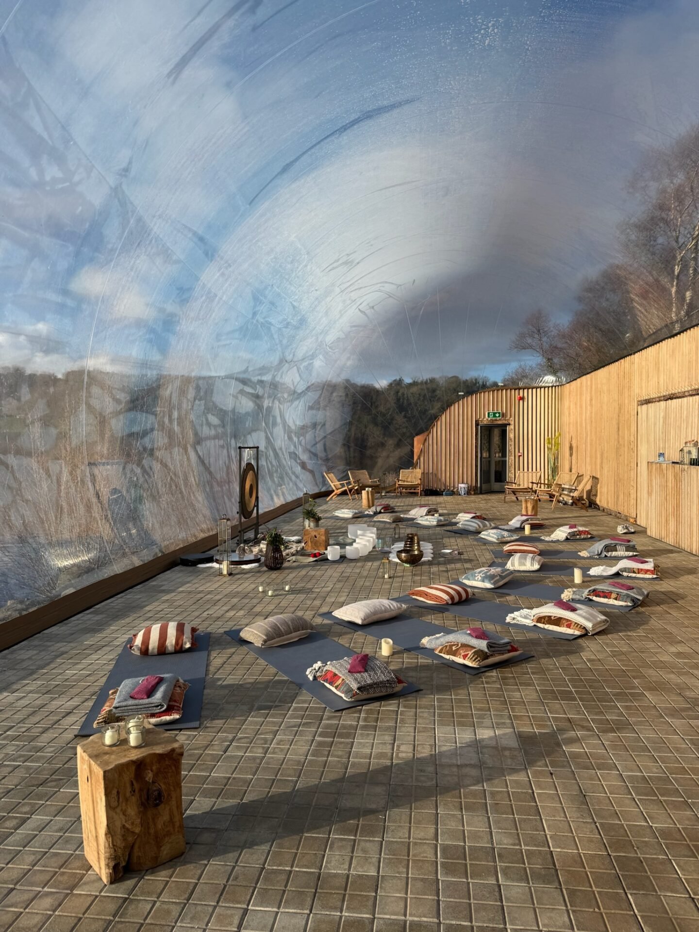 A yoga studio inside a transparent dome with wooden walls, featuring rows of mats, cushions, blankets, and singing bowls on a tiled floor surface