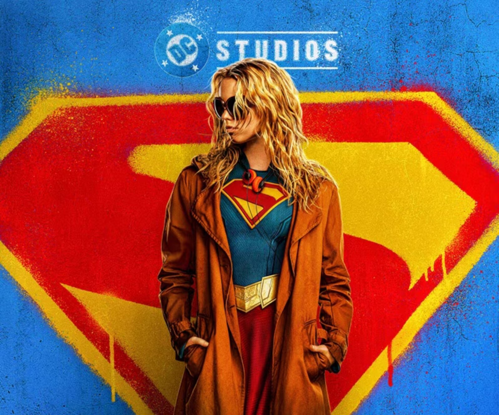 Official Supergirl (2026) poster featuring Milly Alcock as Kara Zor-El wearing her blue and red Supergirl suit beneath a brown trench coat, standing in front of the iconic Superman shield against a blue DC Studios backdrop. - Best Feminist Movies 2026