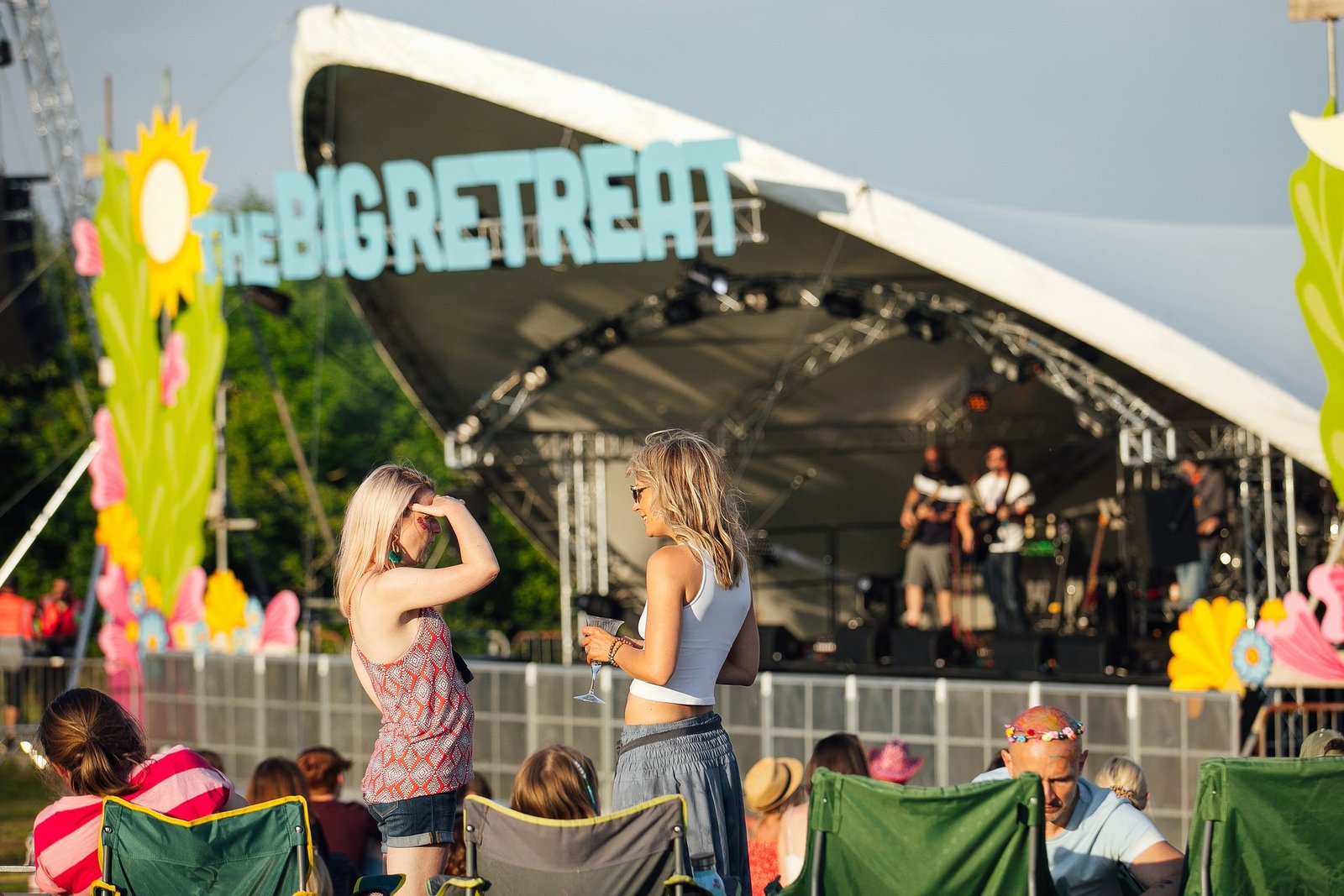 Two women chat in front of a main outdoor stage featuring a live band under a large white canopy at The Big Retreat Festival.