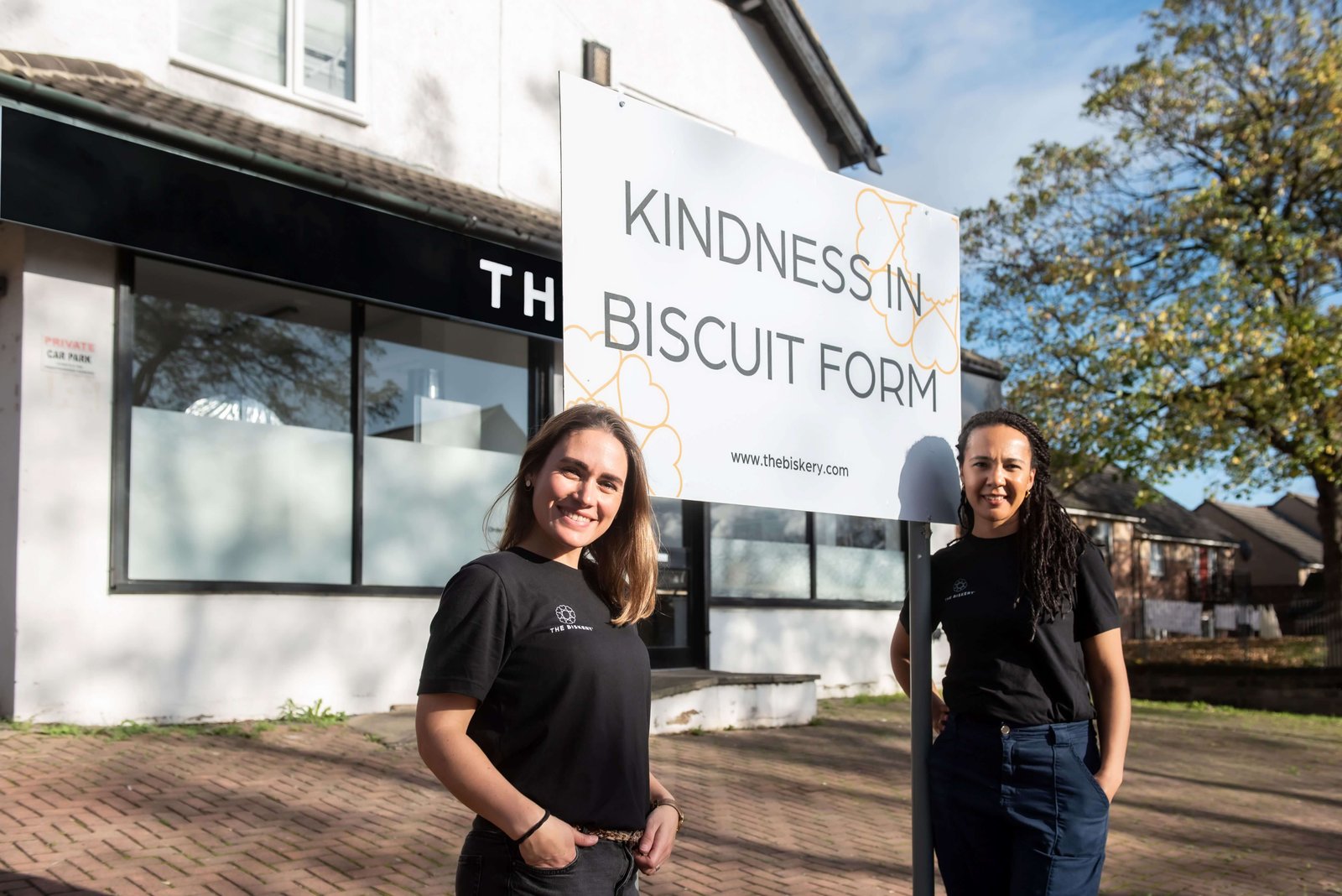 The Biskery co-founders Lisa Shepherd and Saskia Roskam standing outside The Biskery headquarters in Leeds beside a 'Kindness in Biscuit Form' sign