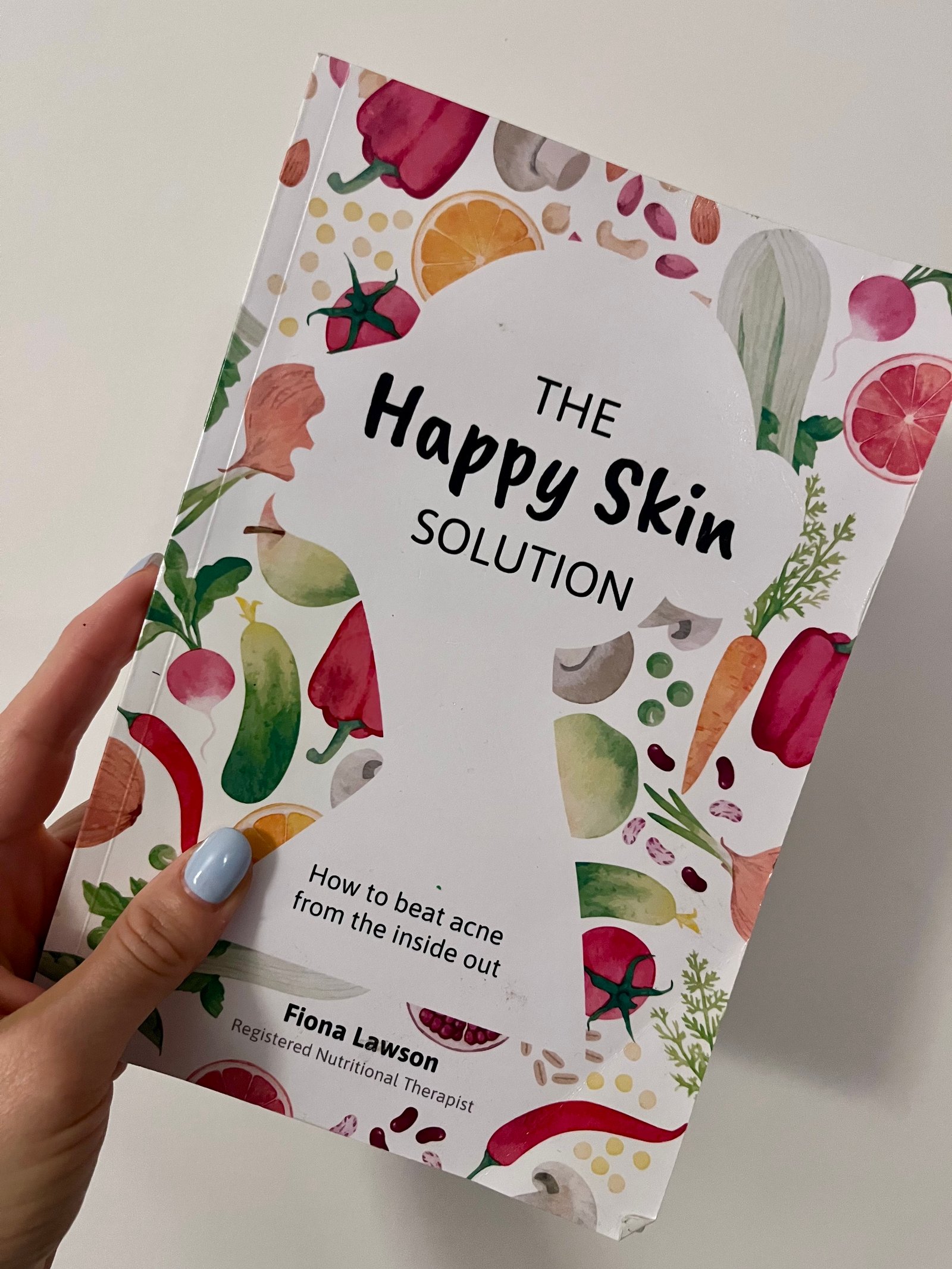 A woman's hand holding up a book called The Happy Skin Solution - How to beat acne from the inside out by Fiona Lawson