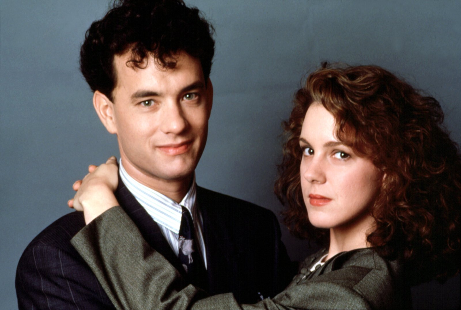 Tom Hanks as Josh Baskin and Elizabeth Perkins as Susan pose closely together in a scene from 'Big', both wearing 1980s business attire against a plain grey background.
