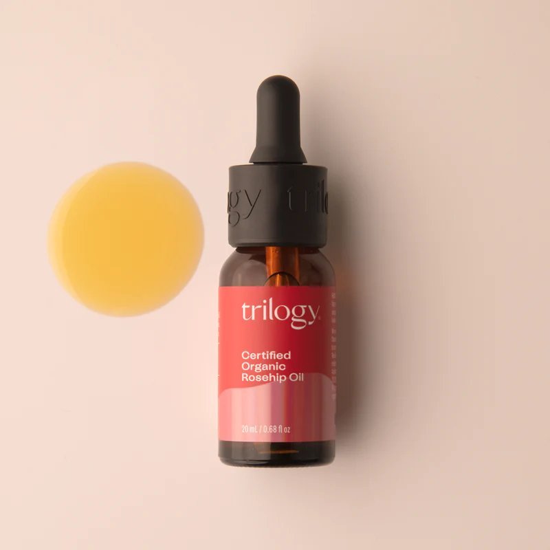 Amber bottle of Trilogy certified organic rosehip oil next to a yellow oil droplet, on a pale background