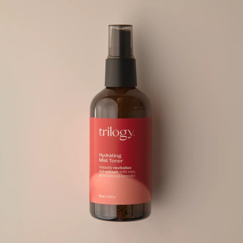 A brown glass bottle of Trilogy Hydrating Mist Toner with a black sprayer, sitting on a light background