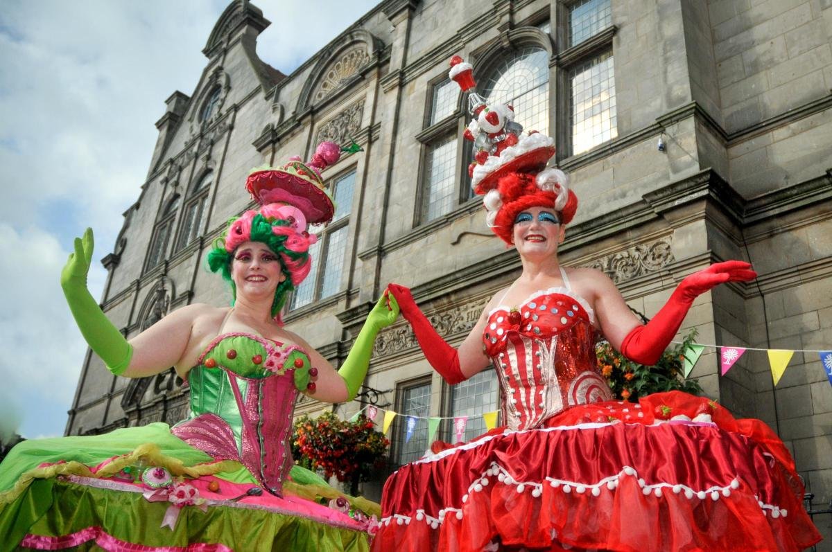 Two performers on stilts hold hands in elaborate, food-inspired costumes—one pink and green, the other strawberry red—complete with towering headpieces. They pose outdoors in front of a historic stone building at the Oswestry Food and Drink Festival.