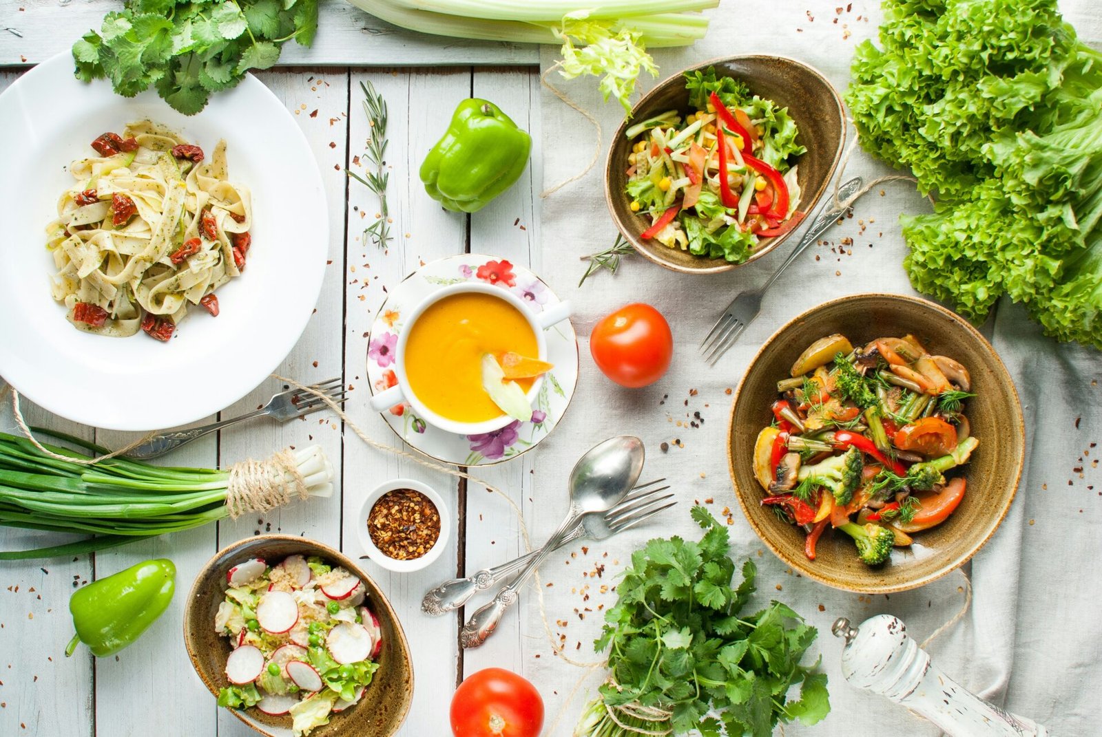 A top-down view of a white wooden table filled with healthy dishes, including pasta with sun-dried tomatoes, orange soup, a fresh salad, and a vegetable stir-fry, surrounded by fresh herbs and ingredients.