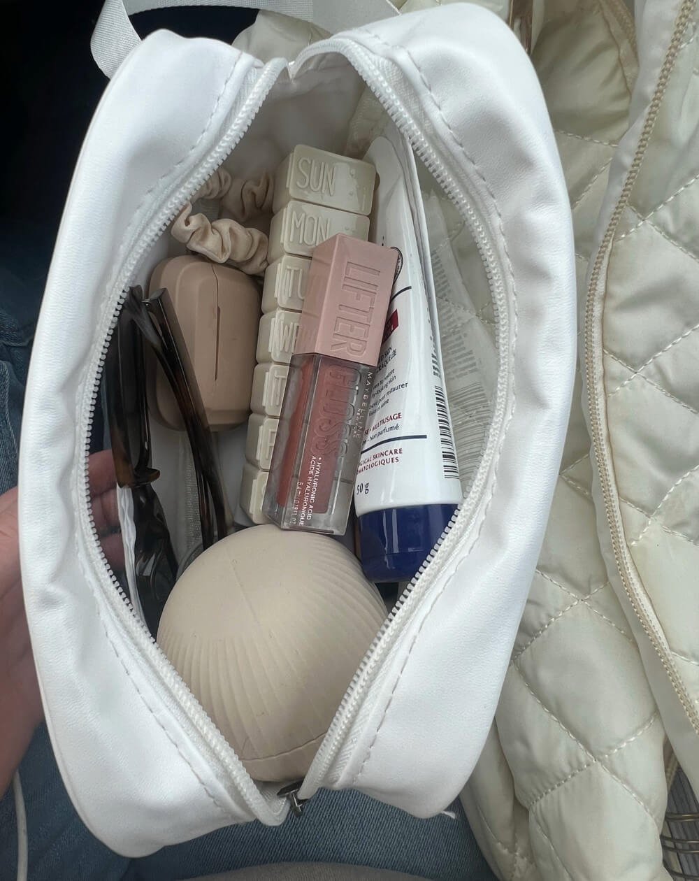 A white makeup bag filled with various cosmetic items, including sunglasses, lip gloss, and a skincare tube