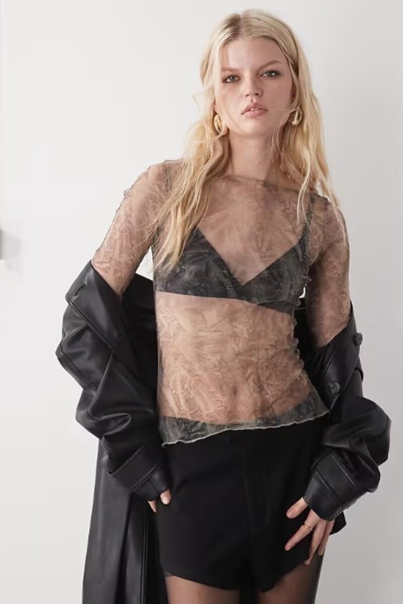 A model wearing Weekday sheer mesh long sleeve top over a black bra, paired with a black leather jacket and shorts