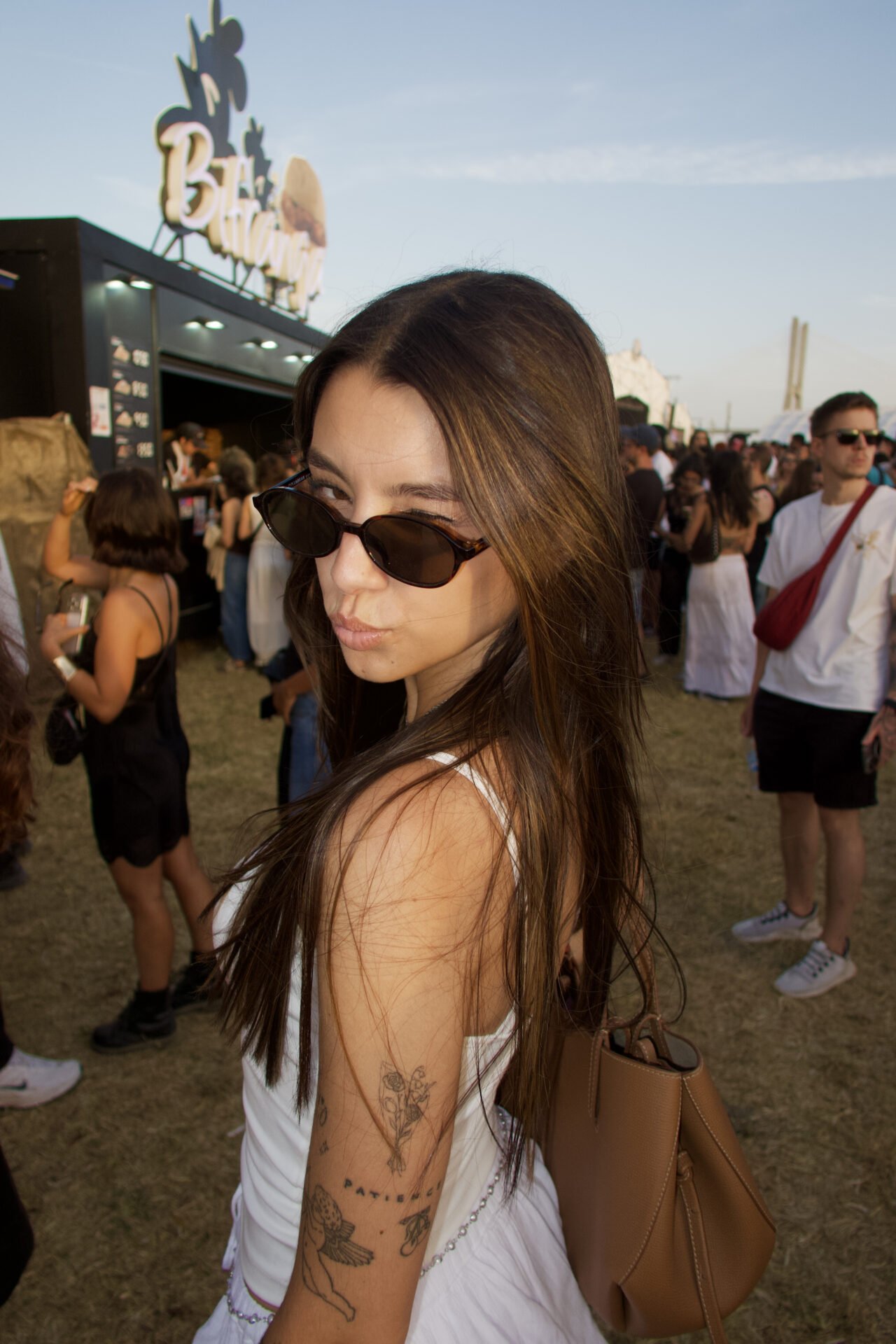 A woman wearing sunglasses and a white festival outfit looks over her shoulder while standing among crowds at a busy outdoor summer festival, with food stalls and festivalgoers in the background.