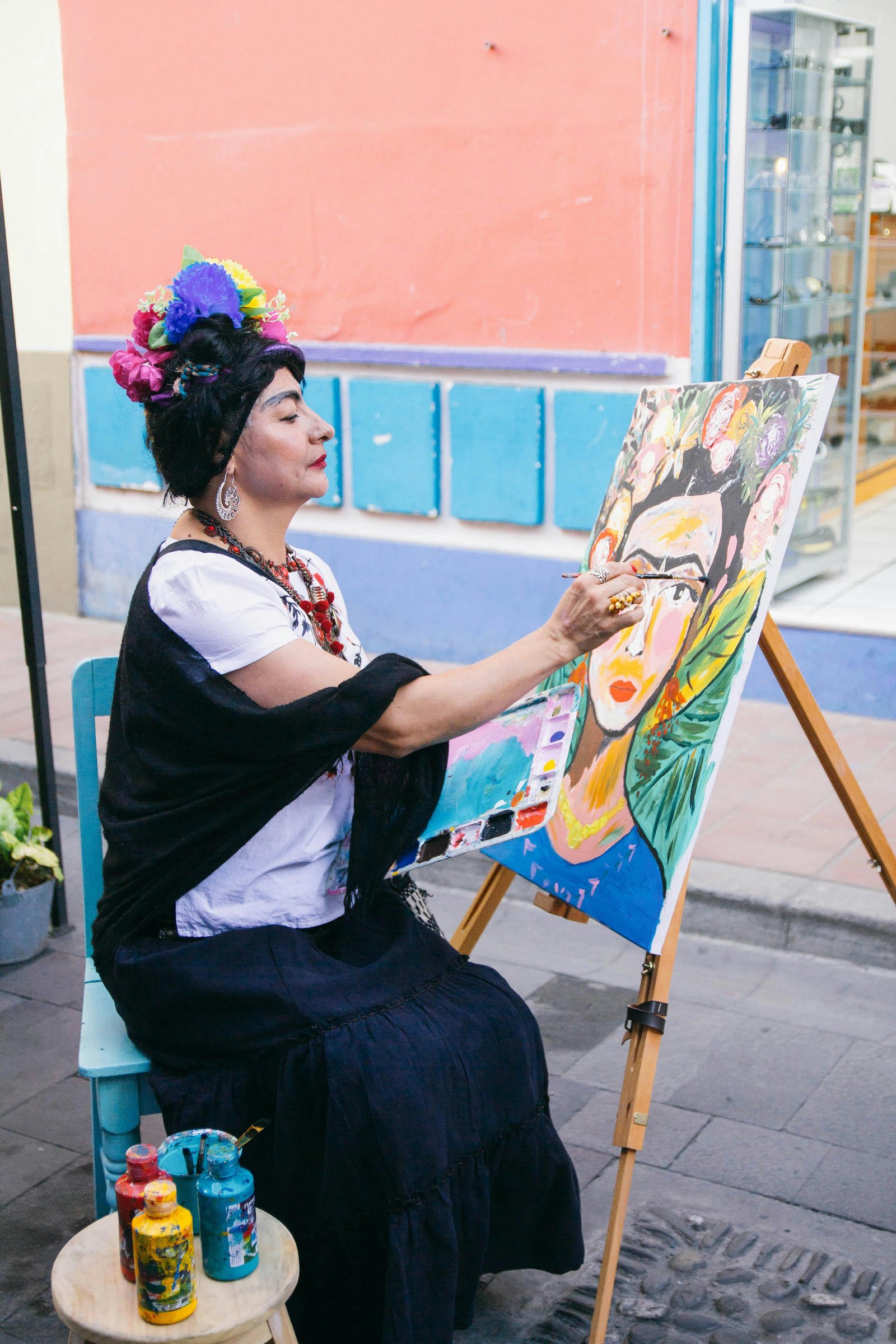 A woman dressed as Frida Kahlo, featuring a floral crown and prominent unibrow, sits outdoors on a blue chair and paints a colorful portrait of Kahlo on an easel.