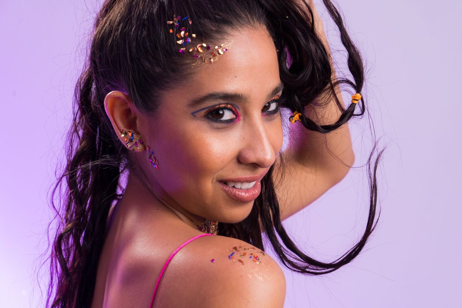 A woman with long, wavy hair adorned with sparkly decorations poses playfully over her shoulder, smiling at the camera against a soft purple background