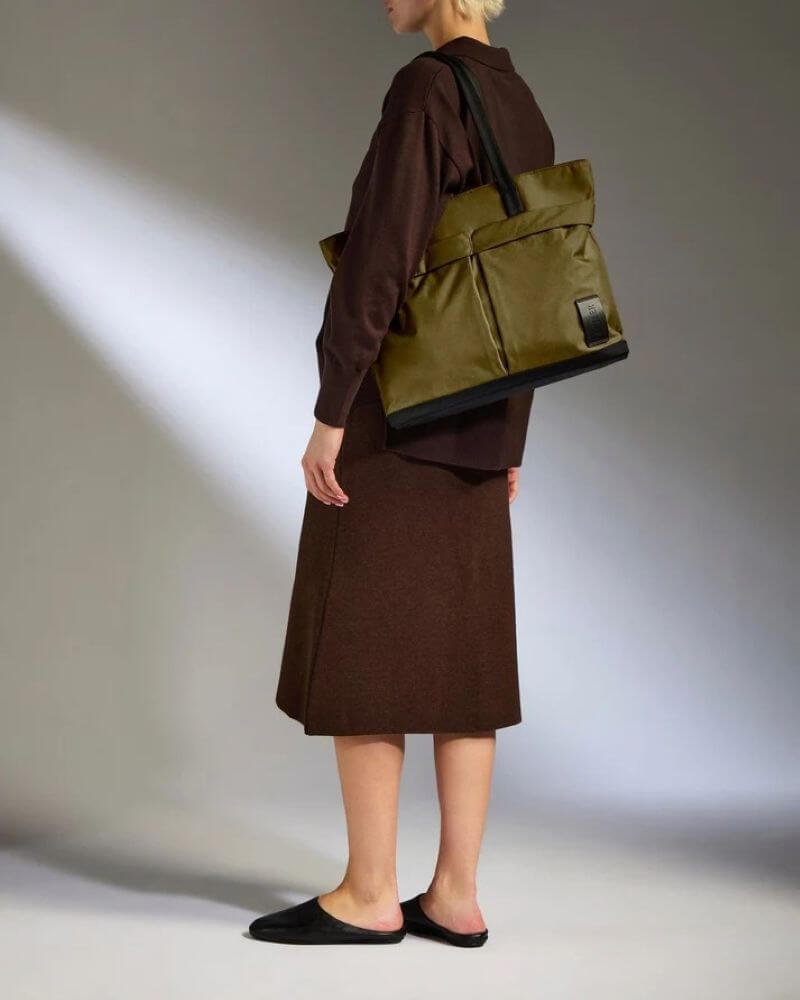 Woman wearing an olive green Antler Urban Core tote bag over her shoulder with black contrast handles. The structured, lightweight tote is styled with a brown outfit to showcase its size and everyday practicality.