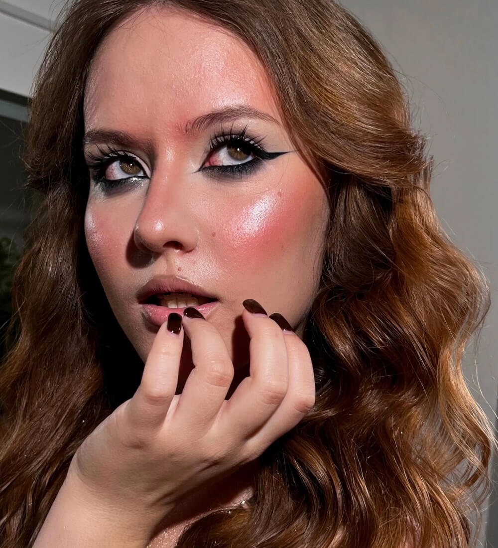 A woman with dramatic Y2K makeup features dark eyeliner and glossy skin, holding her hand to her face